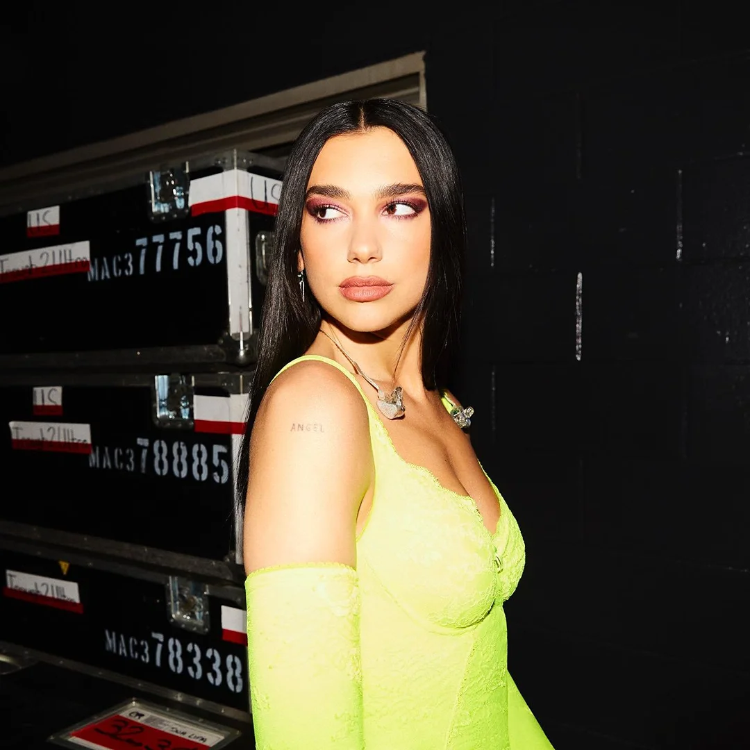 Portrait of Dua Lipa wearing a neon yellow dress with lace and posed in front of a road case with writing on it.