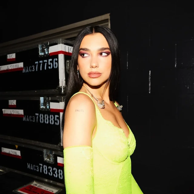 Portrait of Dua Lipa wearing a neon yellow dress with lace and posed in front of a road case with writing on it.