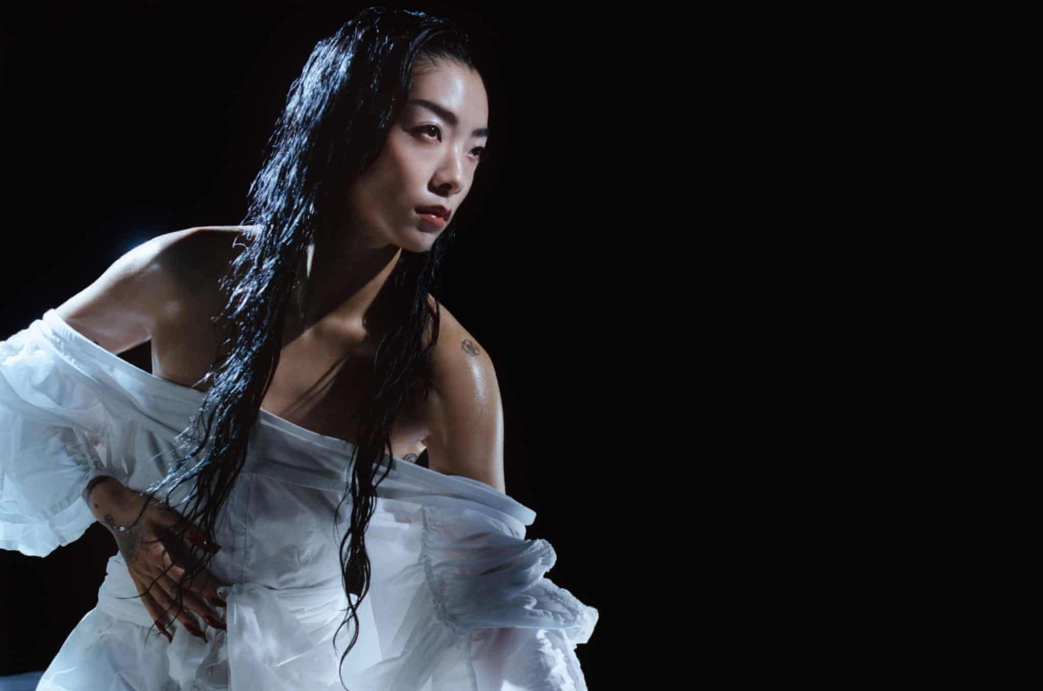 Portrait of an adult female with wet dark hair, wearing a white ruffled garment, in a dark studio setting with focused spotlight.