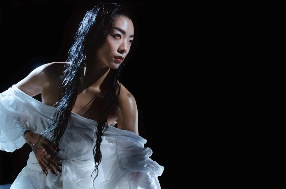 Portrait of an adult female with wet dark hair, wearing a white ruffled garment, in a dark studio setting with focused spotlight.