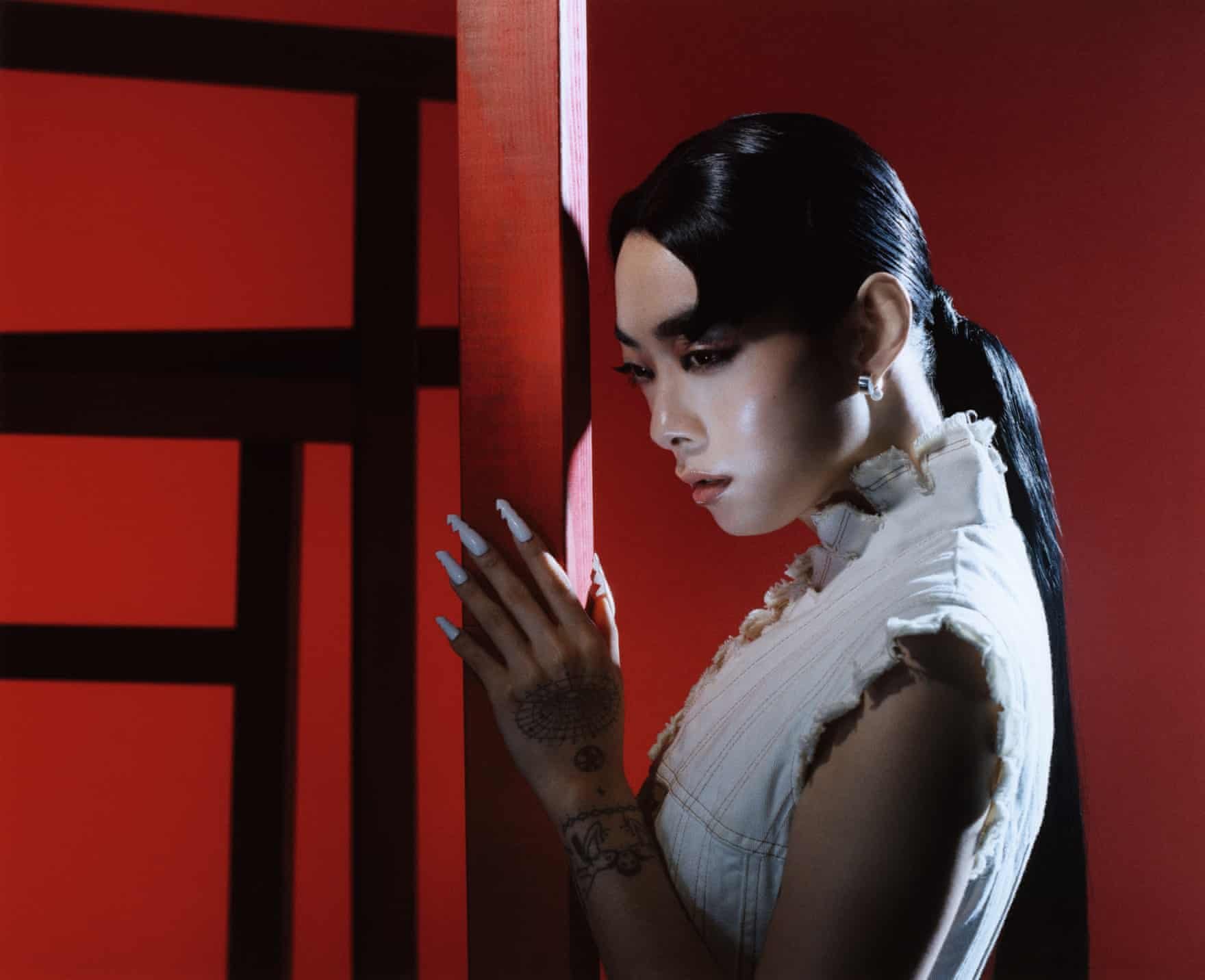 Portrait of an Asian woman in a sleeveless top with long nails posing in front of a red wall with a black square grid pattern.