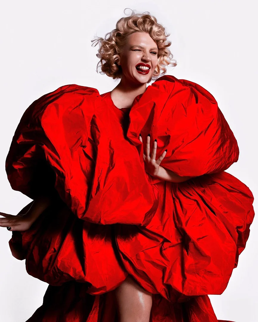An adult female smiles and wears a voluminous red dress with a short blonde wig against a white backdrop, fashion and entertainment shot.