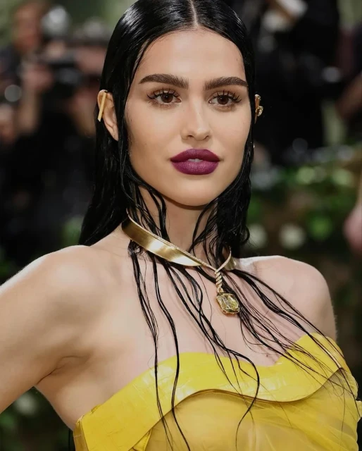 Close-up of a woman with slicked-back black hair, dark lipstick, and a yellow dress, posing in front of a blurred background.
