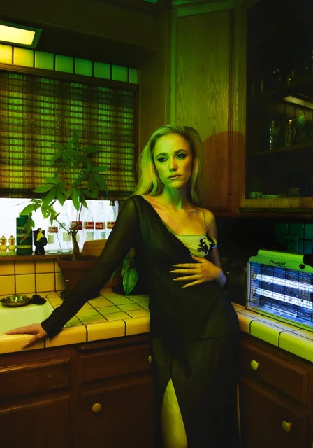 Blonde woman in a black dress leans against a yellow-tiled kitchen counter with dark wood cabinets under green and blue lighting.