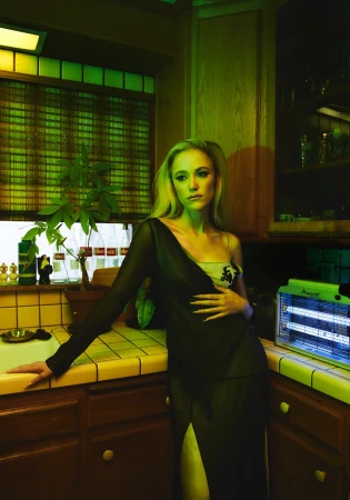 Blonde woman in a black dress leans against a yellow-tiled kitchen counter with dark wood cabinets under green and blue lighting.