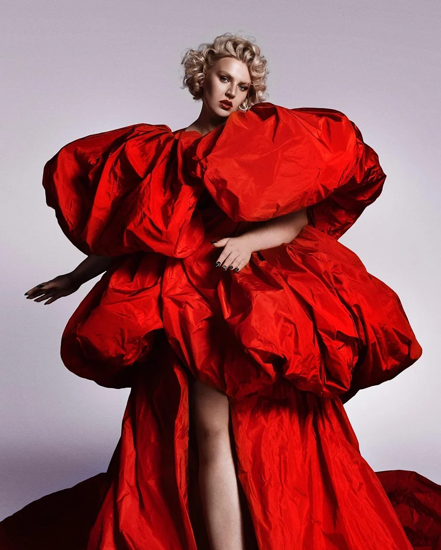 Model with blonde short curls wearing an oversized voluminous red puff dress against a plain neutral backdrop.