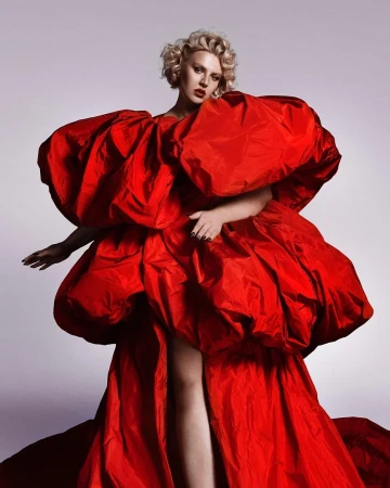 Model with blonde short curls wearing an oversized voluminous red puff dress against a plain neutral backdrop.