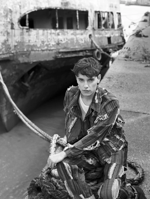 Black and white portrait of a young adult seated on a rope next to a weathered boat, wearing a distressed plaid outfit.
