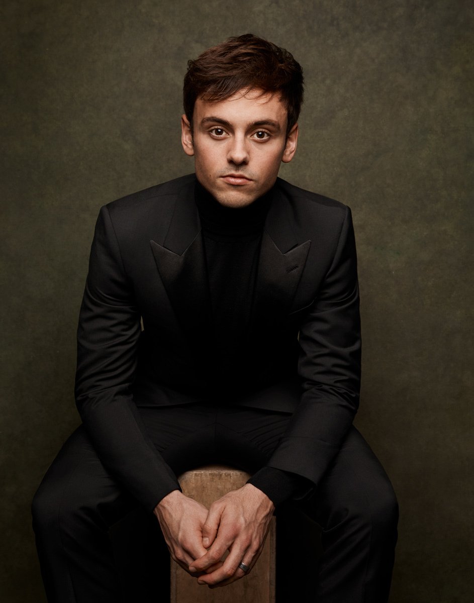 Headshot portrait of Tom Daley in a black suit sitting on a wooden stool against an olive green backdrop in a photo studio. 