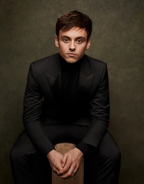 Headshot portrait of Tom Daley in a black suit sitting on a wooden stool against an olive green backdrop in a photo studio. 
