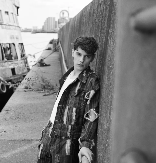 Black and white portrait of a young male model in ripped denim leaning against a wall with a cityscape and boat in the background.