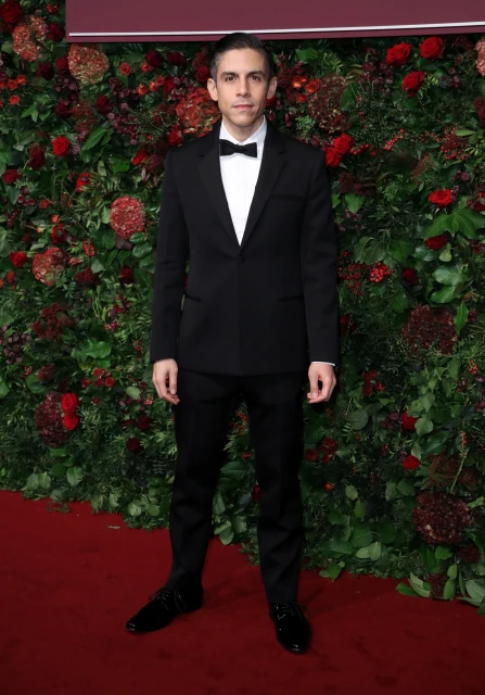 Adult male on a red carpet wearing a black suit and bow tie, standing in front of a leafy green and red flower wall at an event.