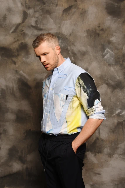 Portrait of a light-skinned adult male with short blonde hair wearing a patterned button-down shirt and dark pants against a textured grey background.