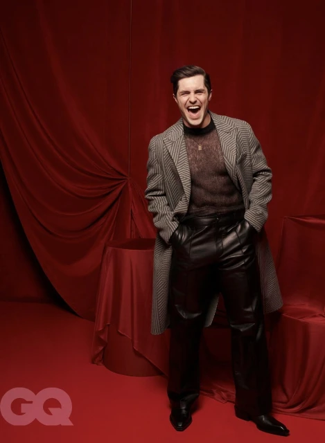 Portrait of a cheerful adult male in a black sheer top and leather pants, wearing a patterned coat, in front of a red backdrop.
