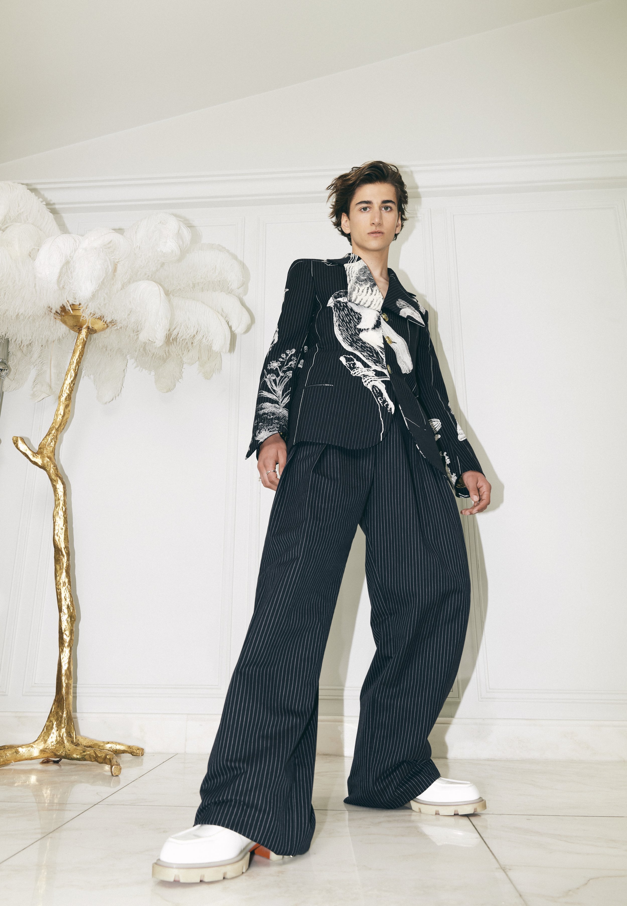 Young male model in a black and white patterned suit and wide-leg striped pants poses in a bright studio with a decorative feather sculpture.