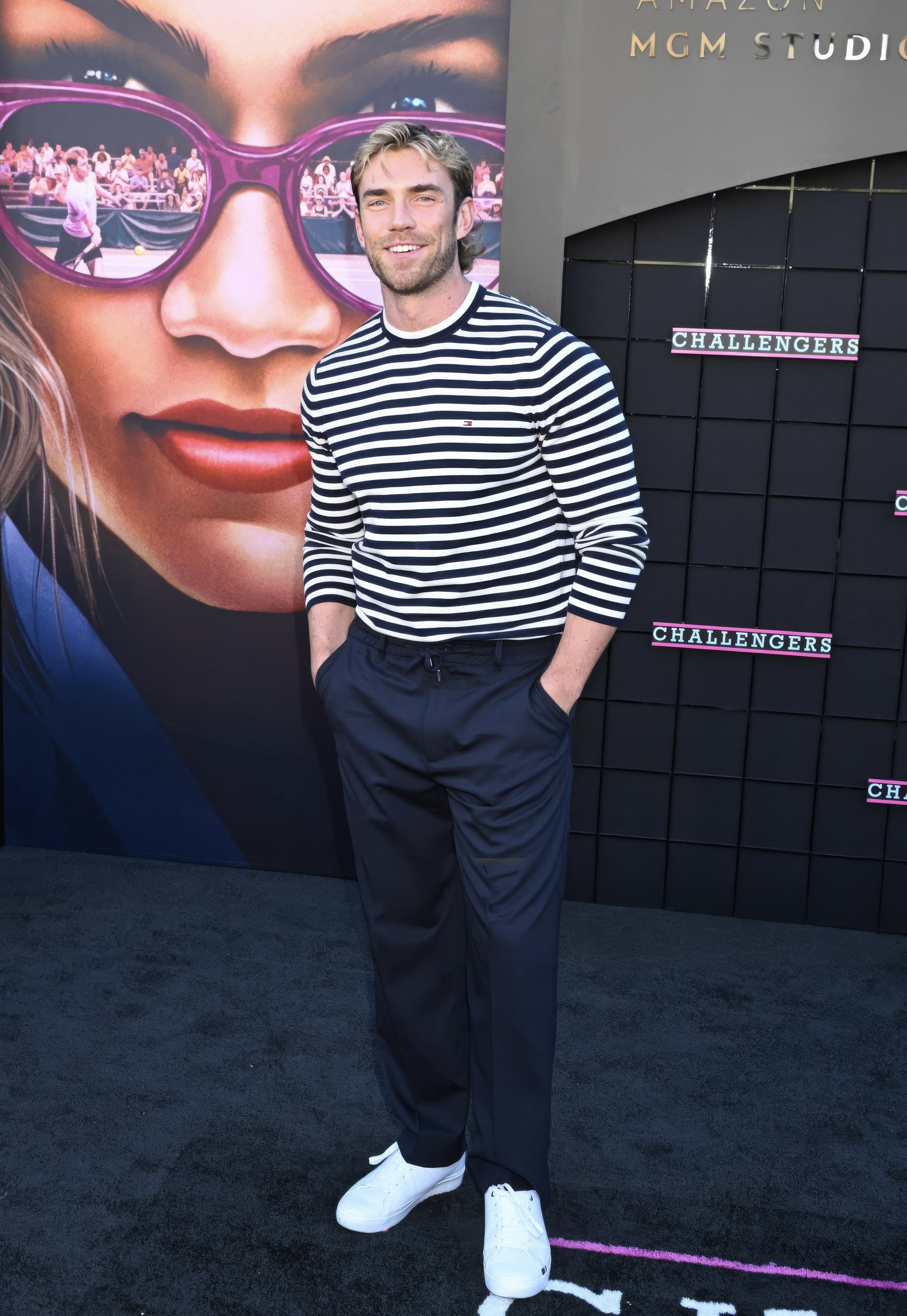 A man poses on a black carpet at the Challengers film premiere event in a horizontally striped long-sleeve shirt, and black pants.