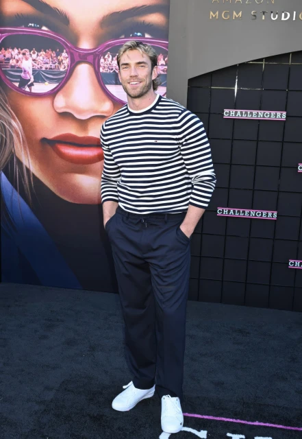 A man poses on a black carpet at the Challengers film premiere event in a horizontally striped long-sleeve shirt, and black pants.