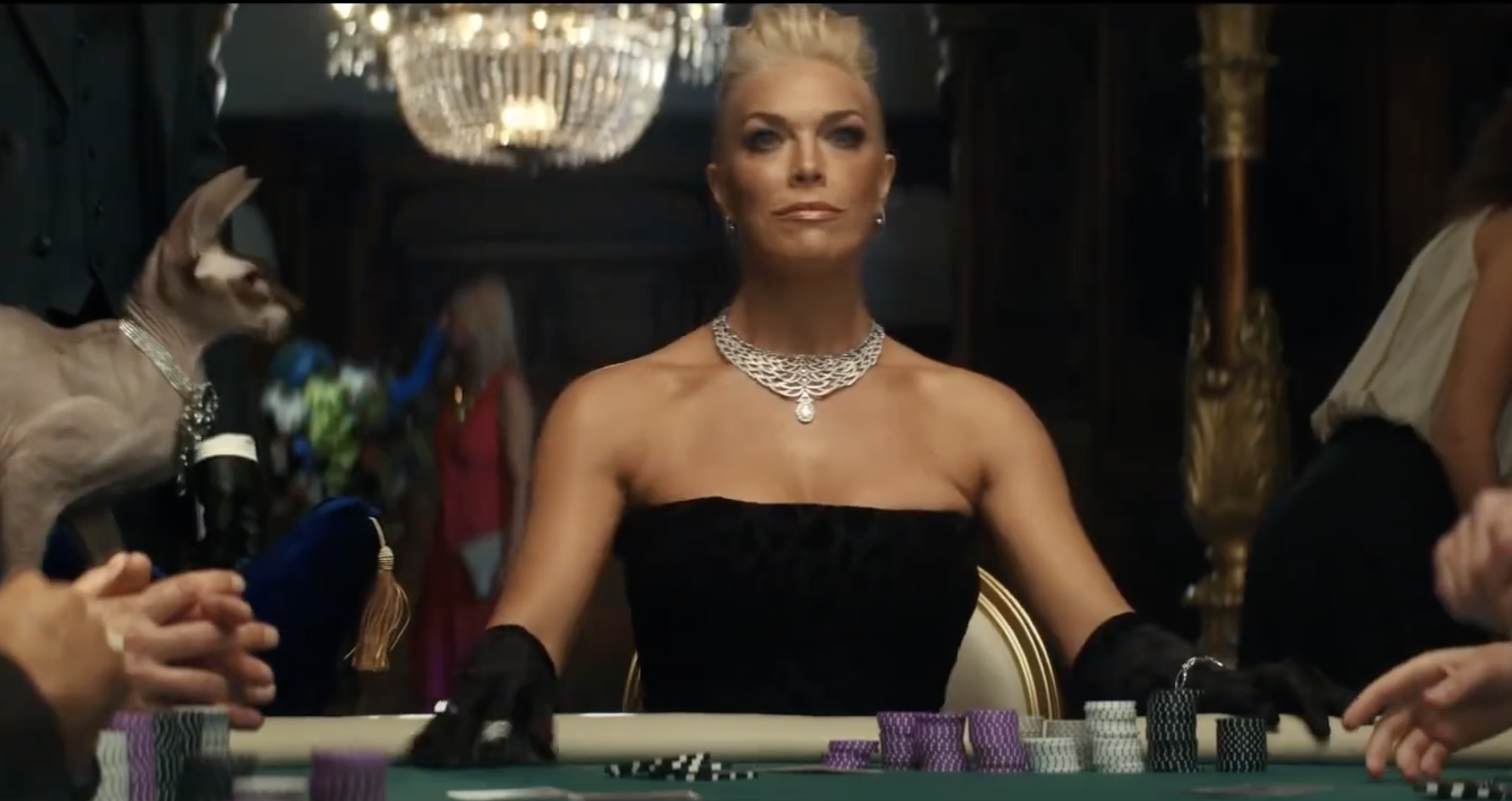 Woman at a poker table wearing a black dress and jewelry, with a hairless cat in the background at an indoor casino setting.