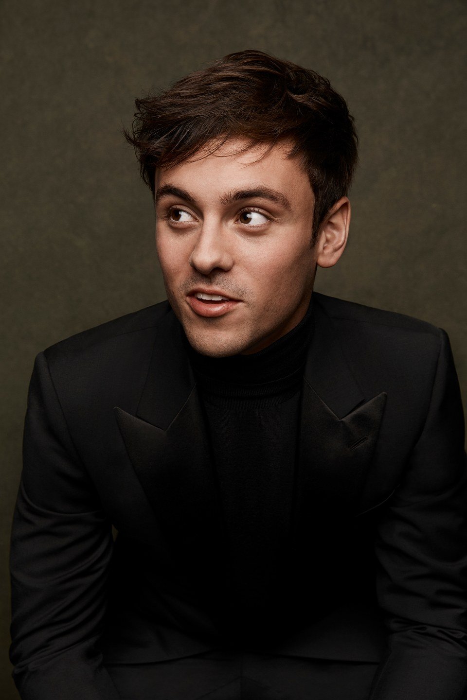 Portrait of a young adult man with short brown hair, wearing a black turtleneck and suit jacket, looking left against a neutral backdrop.
