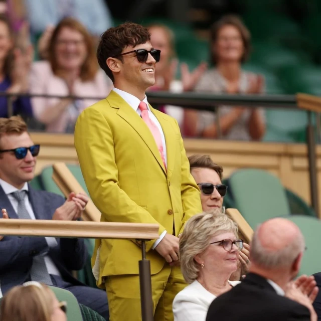 A man in a yellow suit smiles while seated at an outdoor event. People are visible in the background, all enjoying the occasion.