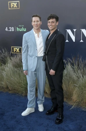 Two adult males pose on a blue carpet at an outdoor event with promotional signage and tall grass in the background on a sunny day.
