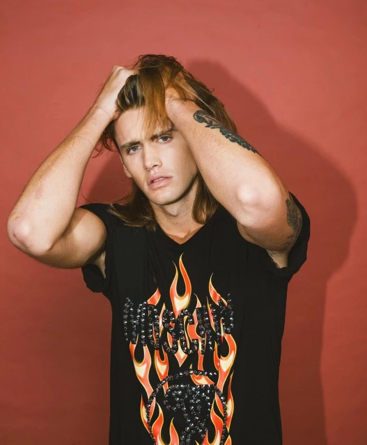Portrait of a young adult male with long hair, wearing a black V-neck t-shirt with flame graphics, against a reddish-brown background.