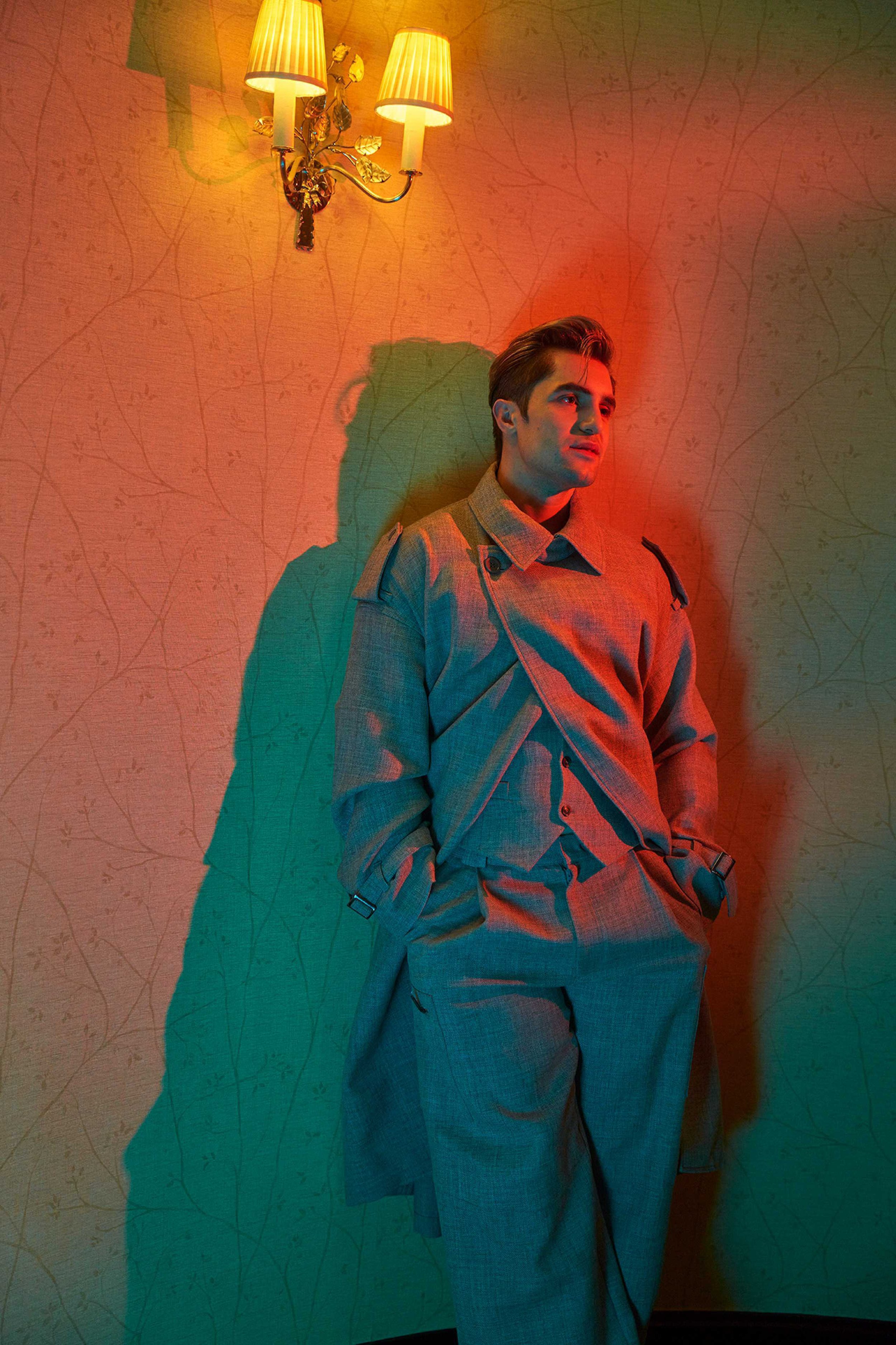 A man in an oversized grey suit stands indoors, leaning against a patterned wall with sconce lighting, casting a teal shadow.