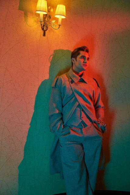 A man in an oversized grey suit stands indoors, leaning against a patterned wall with sconce lighting, casting a teal shadow.