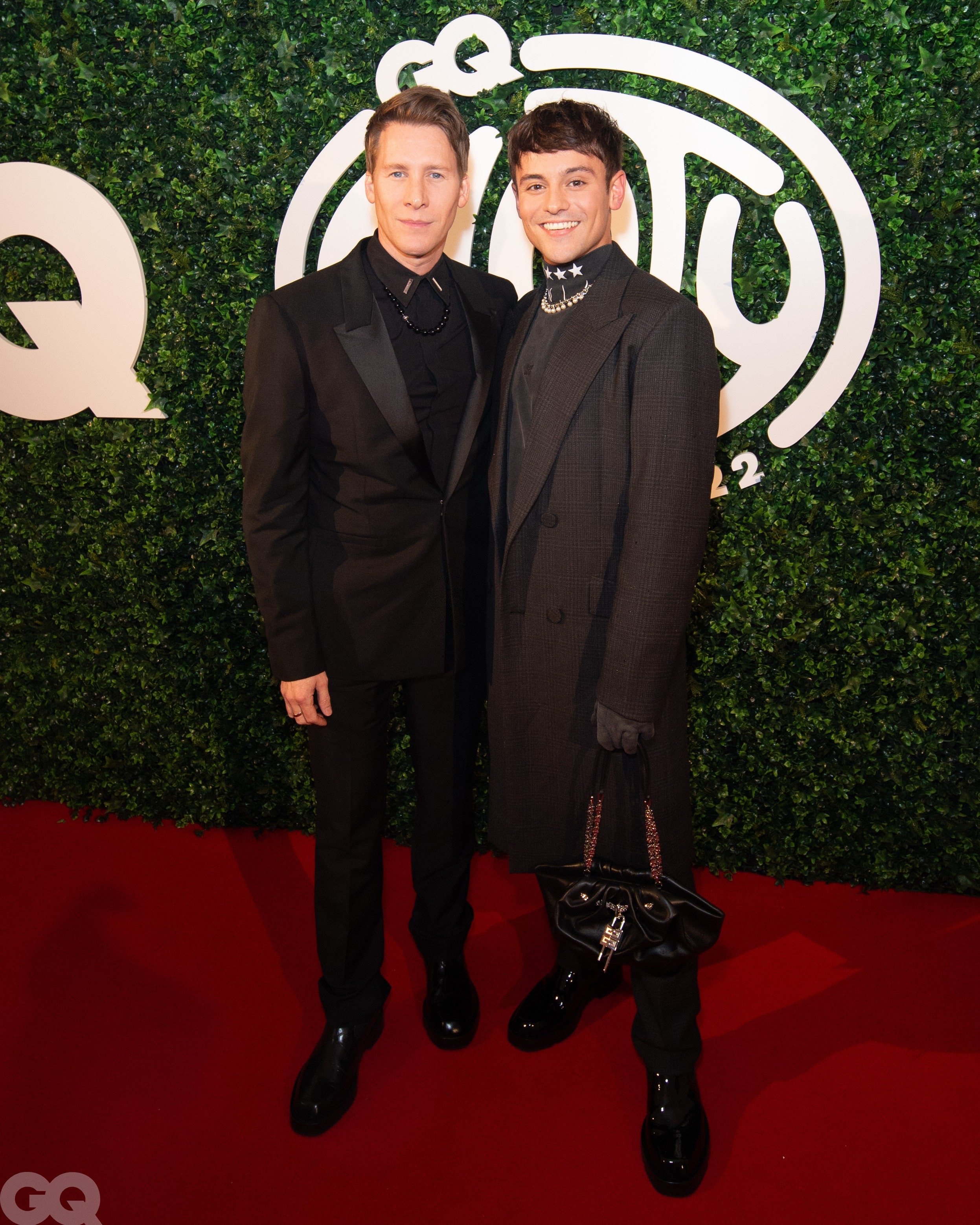 Two men pose on a red carpet in front of a green wall with a white logo. Both are wearing dark suits, and one carries a handbag.