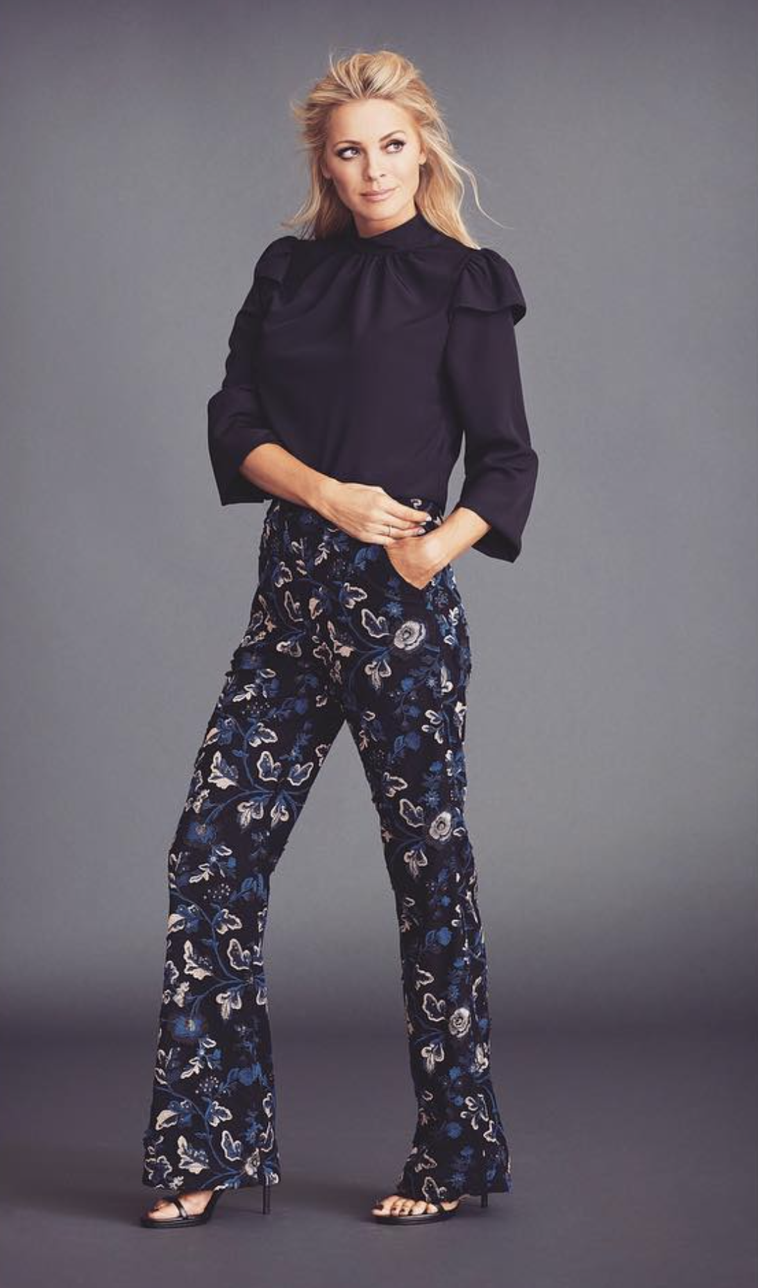 A full-length shot of a poised adult woman in stylish dark floral pants and a black blouse against a solid grey backdrop, fashion pose.