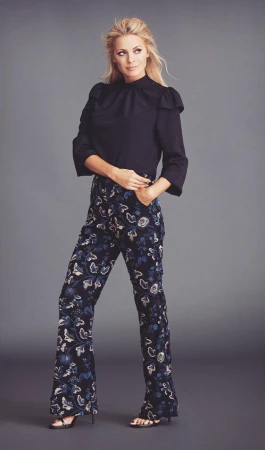 A full-length shot of a poised adult woman in stylish dark floral pants and a black blouse against a solid grey backdrop, fashion pose.