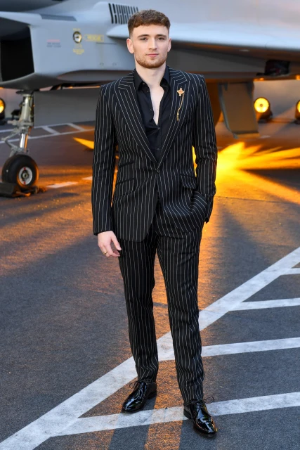 Man in striped suit on tarmac with fighter jet in the background. Adult male poses in formal attire at aviation event, black and white. 
