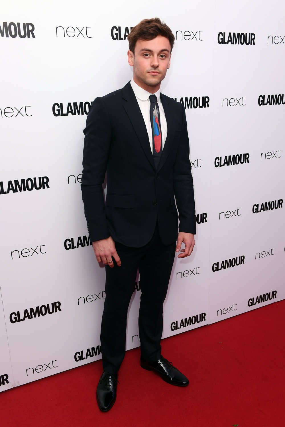Well-dressed man in a suit standing in front of a backdrop with logos. The man is wearing a black suit and tie and black shoes.