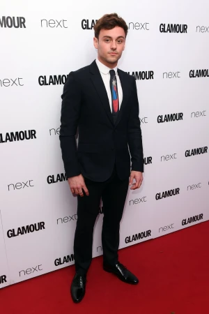 Well-dressed man in a suit standing in front of a backdrop with logos. The man is wearing a black suit and tie and black shoes.