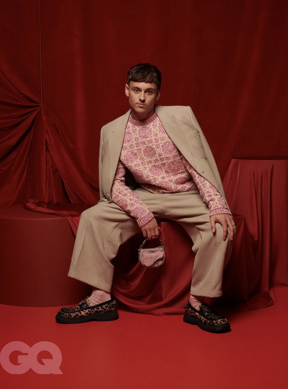 Stylish young man in a patterned pink sweater and beige suit sitting against a red draped backdrop holding a small beaded handbag.