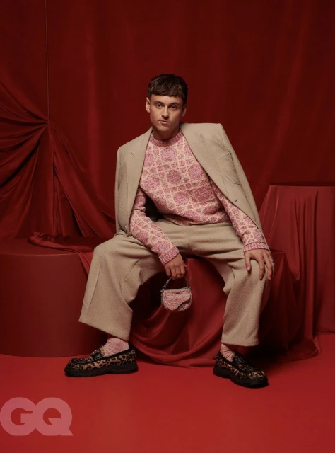 Stylish young man in a patterned pink sweater and beige suit sitting against a red draped backdrop holding a small beaded handbag.