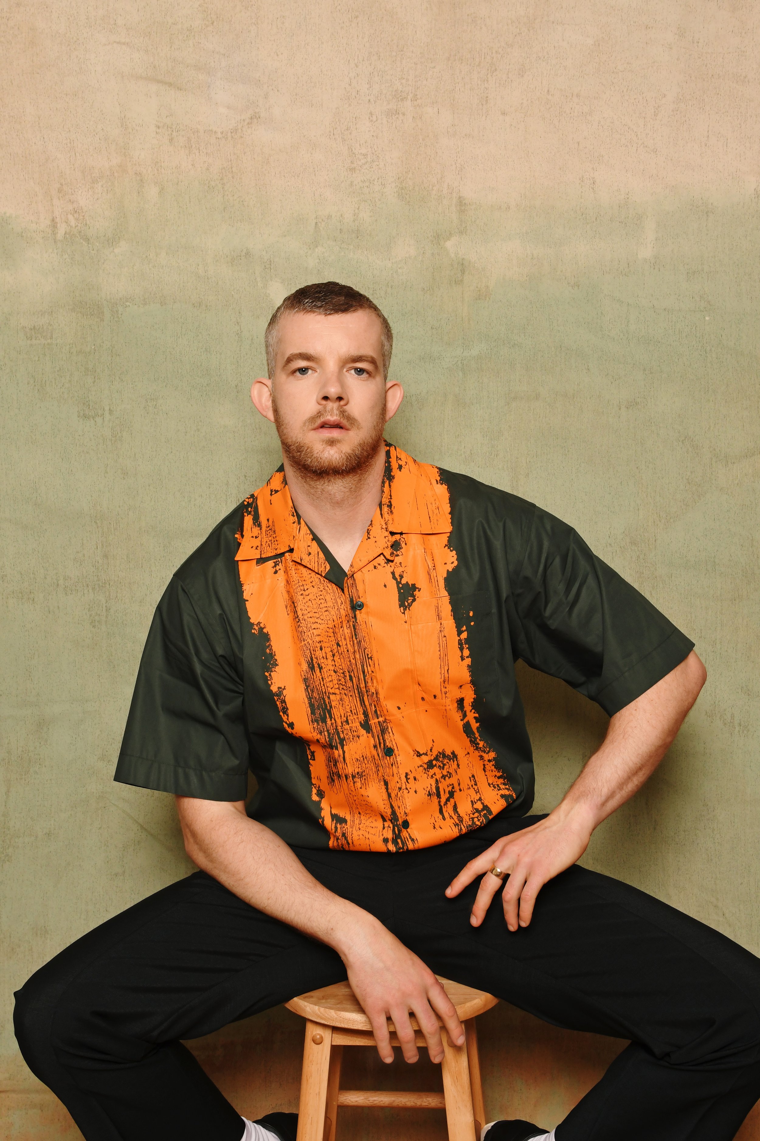 An adult male sits on a wooden stool with a green and orange shirt and black pants against a textured neutral background in a studio.