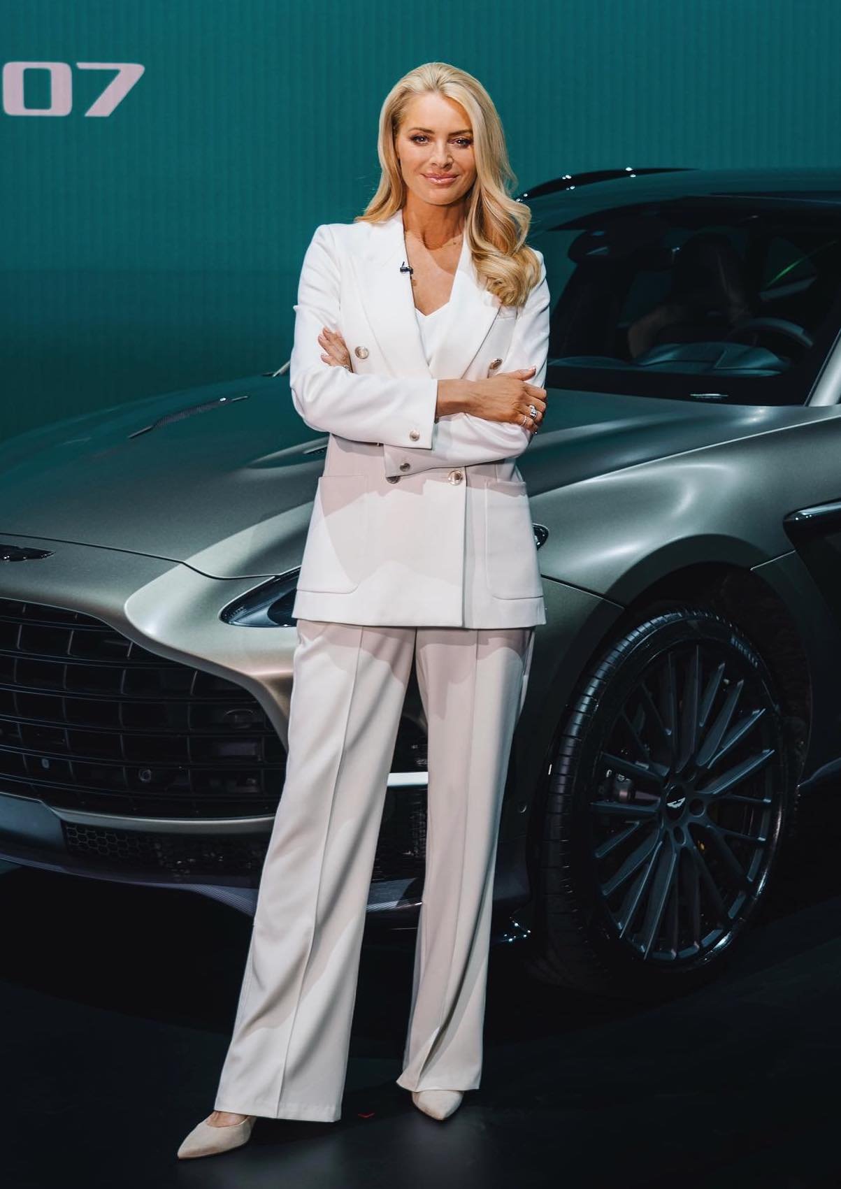 Blonde woman in a white pantsuit stands next to a silver Aston Martin car with a teal-blue background in an indoor setting.