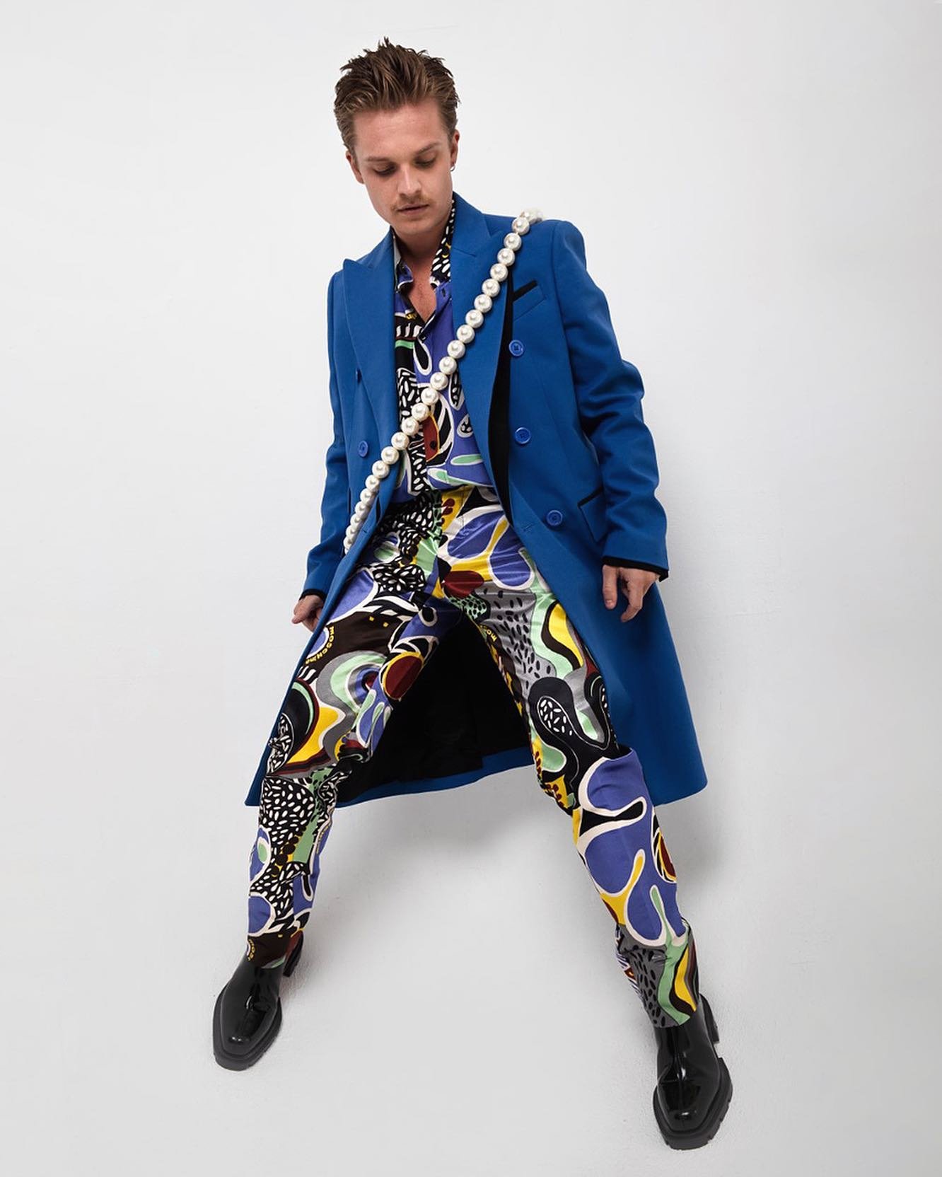 Male fashion model poses in a blue double-breasted coat with patterned pants, a patterned shirt, black shoes, and a pearl necklace.