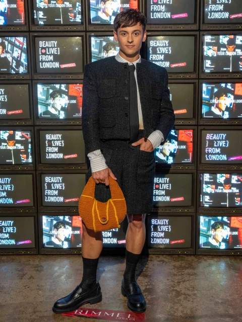 Tom Daley poses in a tweed suit with a handbag against a backdrop of monitors displaying 'Beauty Live From London'.