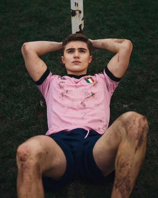 Young adult male soccer player lying on grass with hands behind his head, pink jersey, covered in dirt, outdoor setting, natural lighting.