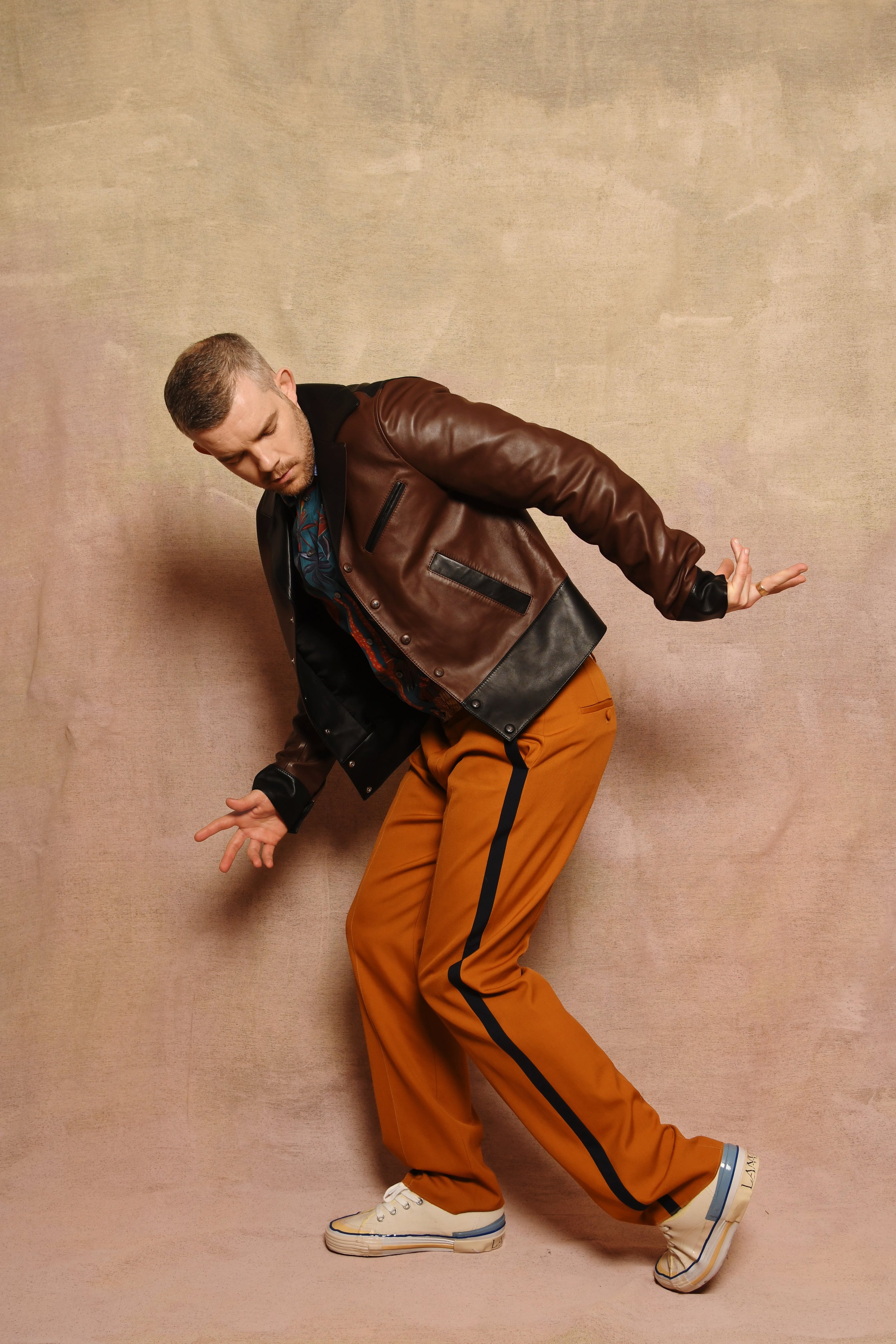 Adult male poses indoors wearing a brown leather jacket and orange trousers with a black stripe, in front of a mottled beige backdrop.