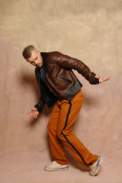 Adult male poses indoors wearing a brown leather jacket and orange trousers with a black stripe, in front of a mottled beige backdrop.