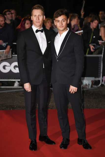 Two adult men in formal black suits pose on a red carpet at an event with a blurred background crowd and signage. 