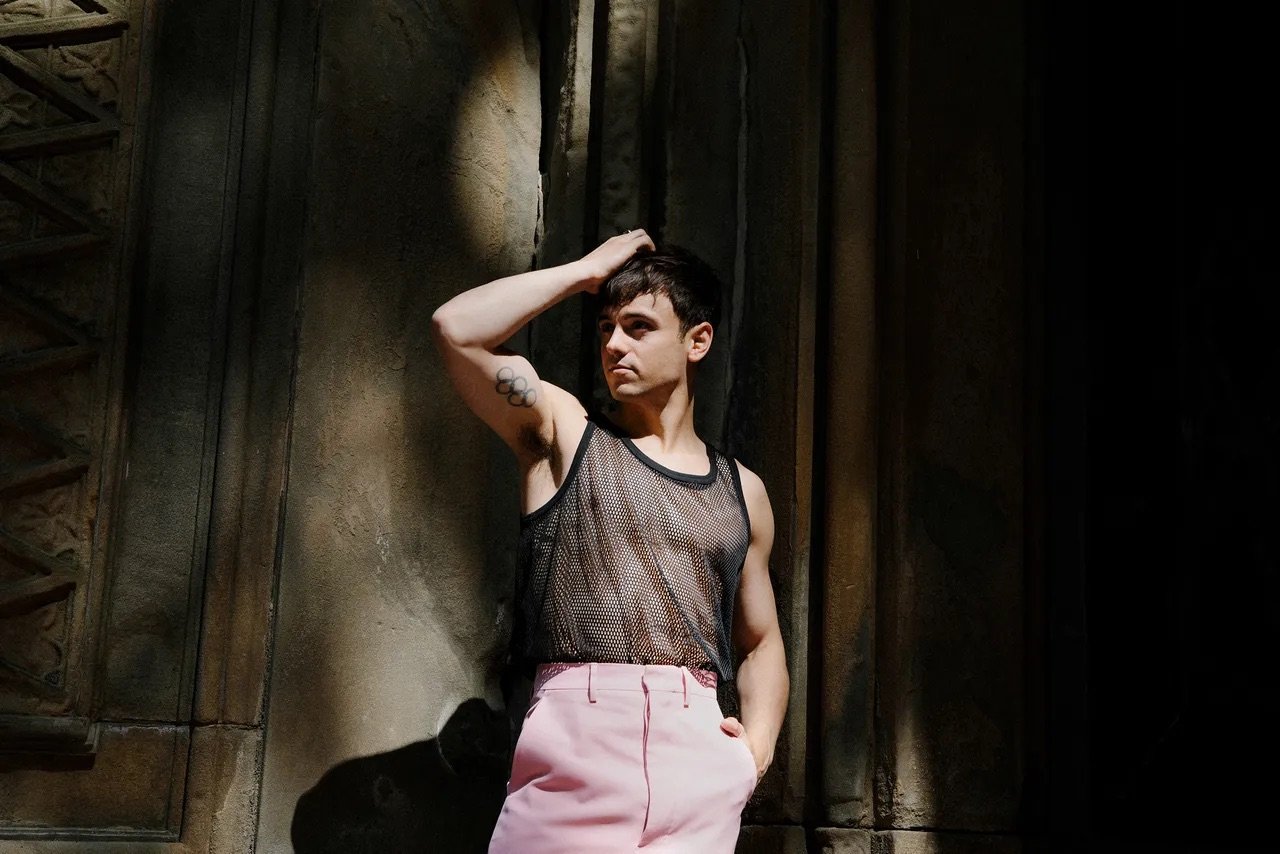 Young adult male poses against a textured wall wearing a black mesh tank top and pink pants in a medium shot with natural lighting.