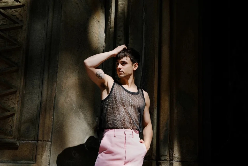 Young adult male poses against a textured wall wearing a black mesh tank top and pink pants in a medium shot with natural lighting.
