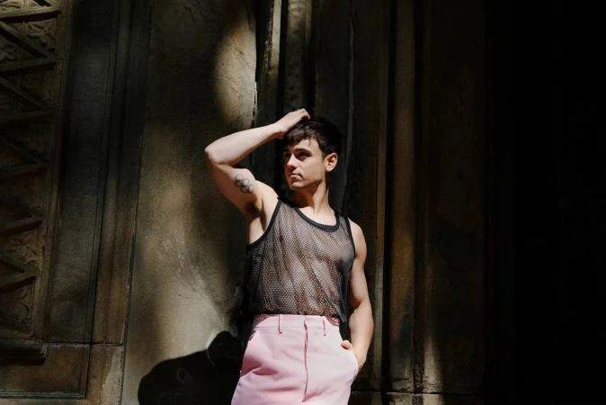 Young adult male poses against a textured wall wearing a black mesh tank top and pink pants in a medium shot with natural lighting.