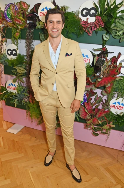 Adult male in tan suit poses against a foliage wall with logos. The man wears an open-collar shirt and loafers, posing in a full shot.
