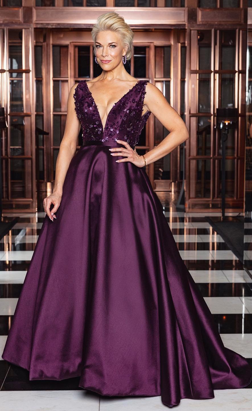Elegant adult woman in a floor-length plum gown with floral bodice, posing indoors in front of a decorative screen.