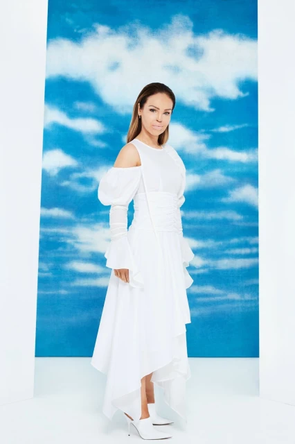 A woman in a white dress with a blue sky and cloud background poses in a fashion shot. The model looks confidently at the camera.